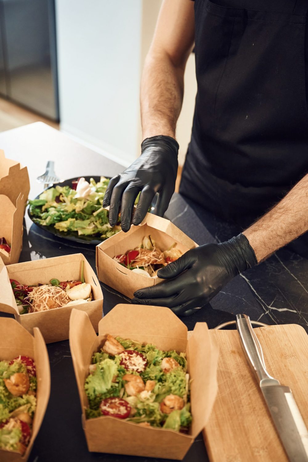 View from the side of meal, vegetables. Man is packing food into the paper eco boxes. Indoors, restaurant.